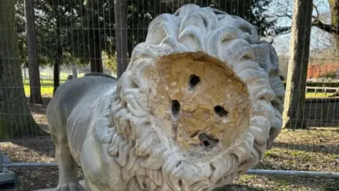 A stone statue of a lion that is missing its face stands in woodland, surrounded by protective fencing.