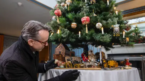 Caroline Sutton Close-up photo of man decorating a table with miniatures characters, carriages and houses, which are underneath a tree 
