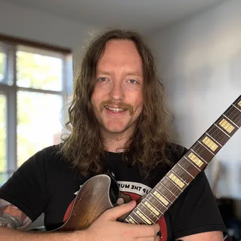 Jamie Donnelly Jamie Donnelly has long brown hair. He is smiling holding a guitar. He has a brown moustache. He is wearing a black t-shirt. He has a tattoo on his arm. There is a window behind him.