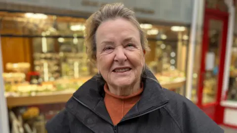 A brown-haired older woman with a black coat and an orange turtleneck. 