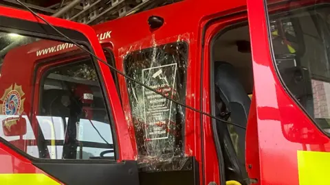 A fire engine pictured from the side with a damaged window covered in clear cling-film type material. The front and back door of the vehicle are open.