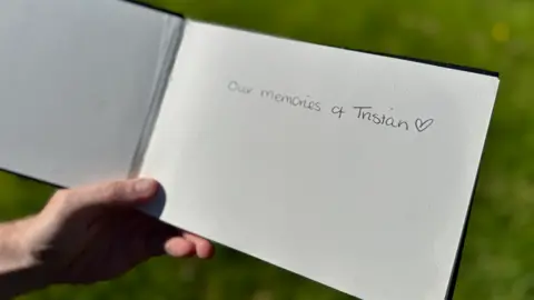 An open book being held with a hand, and green grass behind. In handwriting on the right side, on a blank page it reads "our memories of Tristan" following by a small love heart.