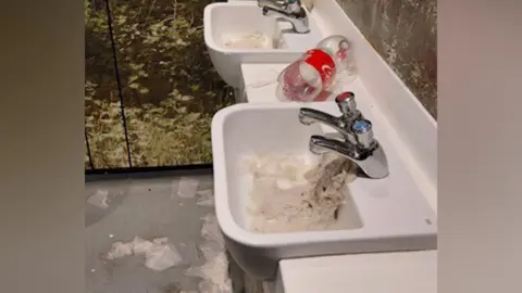 Dartford Borough Council Two basins clogged up with soaked toilet paper. There is an empty coca cola plastic bottle in between the basins. 