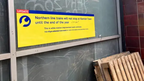 Yellow sign 'Northern line trains will not stop at Kentish Town until the end of the year' with graffiti which says 'which year?'