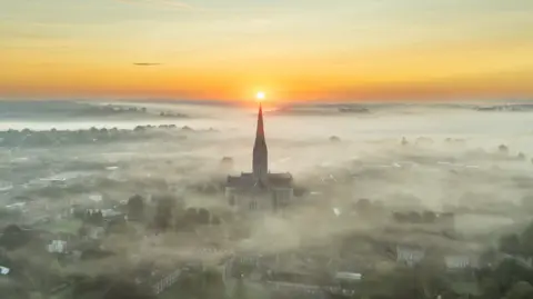 Mike Boss Photography A drone shot of Salisbury Cathedral emerging from fog with the sun just rising on the horizon