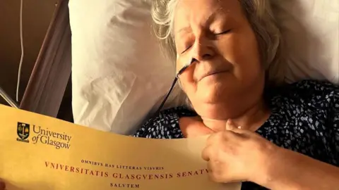 Janey Godley lying in a bed, holding a certificate from the University of Glasgow. She has her eyes closed and is smiling.