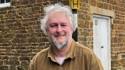 Councillor Gareth Epps has a brown shirt on. He has medium length grey hair and a grey goatee-style beard. Behind him is a road sign.