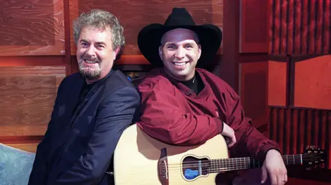 PacemakerPress Two men sitting side by side. One has short grey hair and beard, wearing a black blazer. The other is wearing a cowboy hat and has a red shirt on and a guitar on his lap.