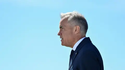 Mark Carney stands in profile against a clear blue sky. His hair is short and grey, and he wears a navy suit and tie and a white short. 