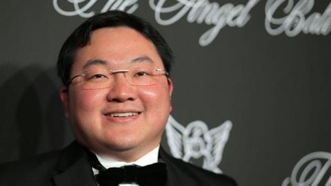 An image showing a smiling Jho Low wearing glasses and black tie, standing in front of a black background