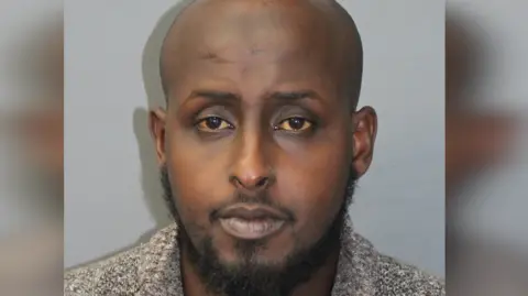 Met Police A adjacent  up   constabulary  changeable  of Mohamed. He is bald with facial hair. Standing against a grey wall. 