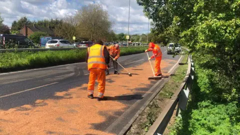 ALEXANDER STEVANOVIC/BBC Three men wearing orange high-visibility jackets are sweeping salt or sand over a dual carriageway using brooms. There are bushes either side of the road.