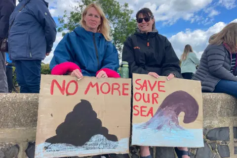 STEPHEN HUNTLEY/BBC Two people sit on a low wall holding hand‑painted cardboard protest signs reading “No more” and “Save our sea”, with other people gathered in the background.