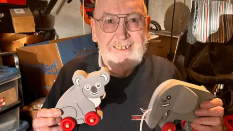 Bill Houghton smiles holding up a wooden koala bear and elephant toy from inside his shed. He has square glasses and a white beard. 
