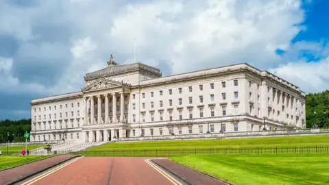 The image shows Parliament Buildings at Stormont on a sunny day. The building is a large, white-stoned, Neoclassical building situated at the top of a hill. There is a road and green hills coming down from it. 