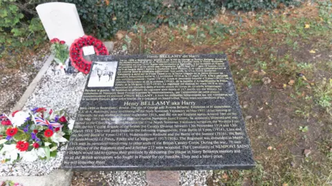 James Clements A dark marble plaque with gold writing, detailing the life of Henry Bellamy in both French and English. It details his service at the Battle of Arras in 1917 and his death. The photo of him on a horse is also on the plaque. Next to it is his grave, which has white pebbles on it, as well as flowers and a poppy wreath.