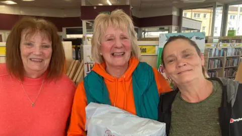 BBC Three women stand together smiling in front of bookshelves in a library