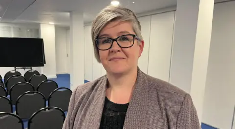 Deputy Haley Camp - A woman with grey hair, wearing black rimmed glasses. She has a grey checked jacket on over a black blouse. She is standing in front of rows of chairs, organised for a presentation.
Copyright