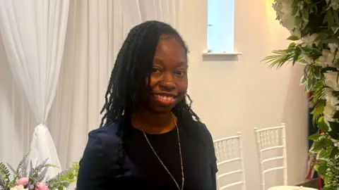 A woman wearing a black dress, gold pendant necklace and gold nose stud smiling into camera. She is standing in front of a white voile curtain with pink and purple flowers arranged in a vase and white chairs are to her right.