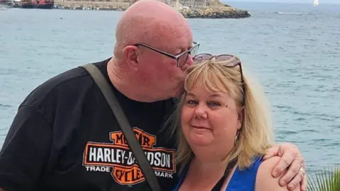 Family handout Sid and Donna stand together on a seaside promenade with a calm sea behind them. Sid is wearing a black T‑shirt with a visible “Harley‑Davidson” logo and has is arm around Donna. He is kissing her on the side of her head. Donna wears a blue sleeveless top and has sunglasses resting on her head. In the background, there is a rocky harbour or breakwater with boats moored nearby and a small sailboat visible further out on the water.