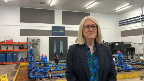 Baroness Jacqui Smith with shoulder length blonde hair and a navy suit and patterned green and blue shirt stands in front of the blue pipes used for training apprentices at Severn Trent's Academy.