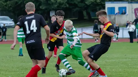O'Neills Foyle Cup A young footballer in green and white strip is surronded by opponents on the field of play. he has the ball at his feet, as he is being tackled. the opponents are in a black strip