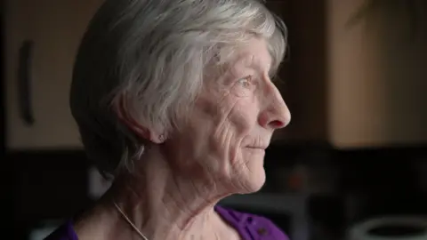 Close up of an elderly lady looking out of a kitchen window with a serious expression on her face. She has short grey hair and is wearing a knitted purple top.