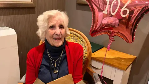 A woman with white hair in a red cardigan and black polo neck. She is wearing gold necklaces and has a helium balloon with 106 on it.
