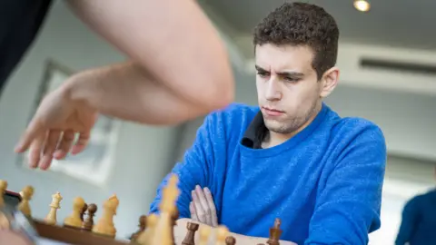 Getty Images Naroditsky sits in front of a chess board with his arms crossed. He is wearing a navy shirt and a blue jumper.