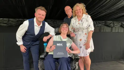 Four people on a stage with one of them in a powered wheelchair holding a National Star College Record of Achievement folder. Three other people stand around the wheelchair user, with one person leaning forward beside the chair and two standing behind. 