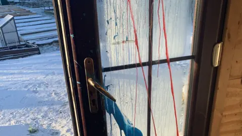 Yvonne Allen Picture of the door to the communal cabin where paint has been thrown up against it. The allotment, covered in snow, can be seen outside.