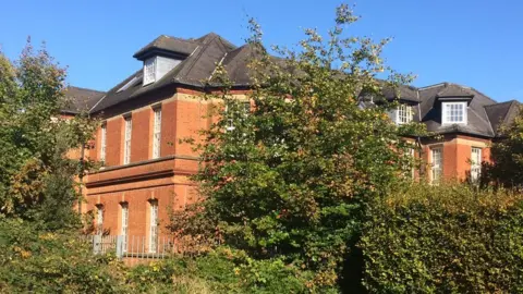 A large red brick building with many windows pictured behind a fence and trees.