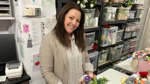 A woman is standing behind a desk that has brightly coloured flowers on it. She has dark brown hair and is smiling at the camera. She is wearing a white and beige striped top with a beige blazer. Behind her are boxes full of flowers. 