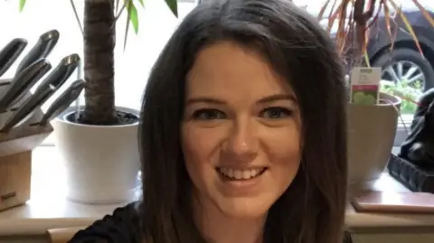 A woman with long, dark, straight hair who is looking ahead and smiling. Behind her are a set of knives in a wooden knife block and two potted plants on a windowsill. 