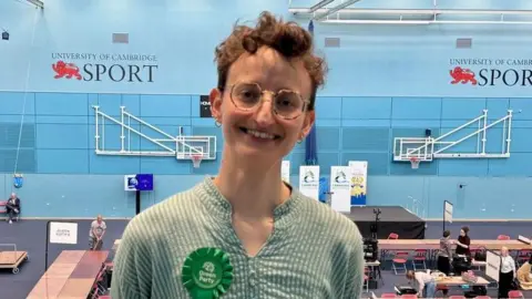 Matt Webb/BBC Kathryn Fisher looks into camera with a sports hall behind her. Kathryn has short brunette hair, rounded glasses ad is wearing a green top with a Green Party rosette.