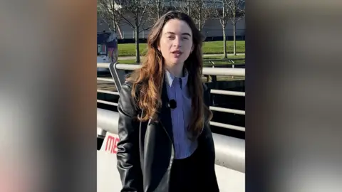 A young woman with brown hair walks on a bridge that has grey railings. It is a sunny day and she wears a blue striped shirt and black leather coat.