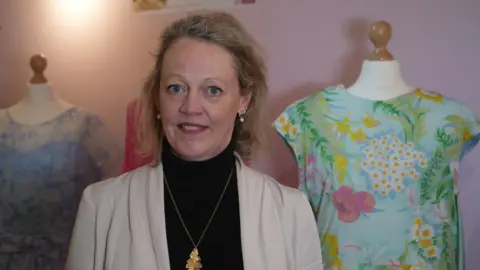 Nicola Haseler Tara Parker pictured in front of some of Barbara cartland's dresses on display. She wears a black polo neck jumper with a cream jacket open, a gold necklace of an oak leaf and several earrings. She has blonde/grey hair half scraped  back and is smiling at the camera.