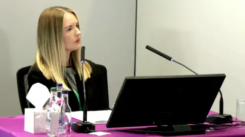 Lampard Inquiry Service manager Chloe Cawston sits in front of a table at the Lampard Inquiry. She has long blonde hair and is wearing a black jacket and a grey top underneath. There are two microphones pointing towards her and a laptop in front. To each side there are bottles of still and sparkling water and a box of tissues.