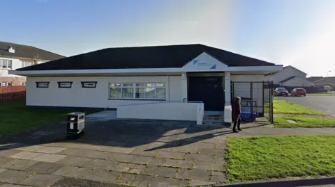 Google A picture of the outside West Winds community centre. It is a single storey building with a sign above the door, a window and a big door. There is a person walking outside the front and a bin on the path out the front.
