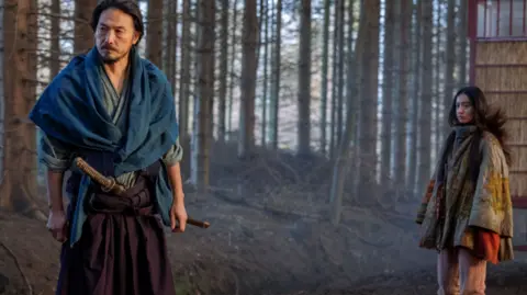 Lionsgate A man with a dark hair and a beard wearing a traditional samurai blue outfit with a sword in his waistband stands next to a woman with long black hair wearing native american clothes 