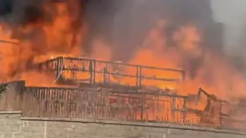 A burning fence and the frame of a house is all that is visible in this picture from  below a wall of orange flames. Black smoke is coming up from a bungalow above a grey brick wall. 