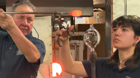 BBC/Jim Scott An older man and a younger woman are making art work or a glass bulb-like item. They are holding it with special tongs. A red hot poker is held by the man. There is a light in the background.
