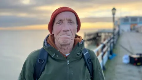 George Carden / BBC Fisherman Bas standing with his rods behind him smiling at the camera wearing a jacket and beanie 