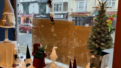 A Christmas scene with a Christmas tree on the right hand side and other Christmas characters behind the broken window pane, which is being protected by bits of cardboard