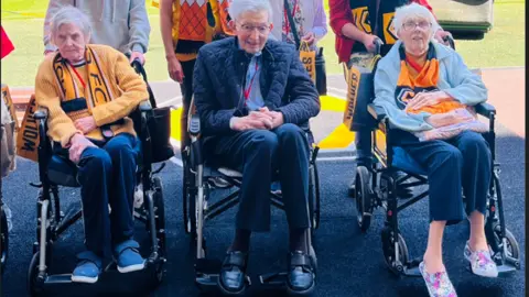 Ashberry Healthcare Three residents sit in wheelchairs with Wolves scarves on by the pitch. Some other people stand behind them.