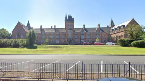 A large brick building sits on top of a lawn of green grass. The sky above is clear and blue. At the bottom of the frame is small black iron fencing. In front of the building there are two red cars and a white car. 