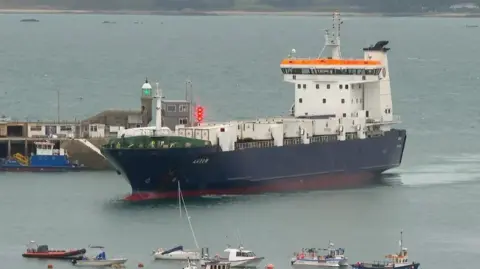 A large red, white and blue vessel travels along calm waters into a harbour area. Smaller boats are moored on buoys in front of the large boat. 