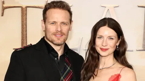 Getty Images A red carpet premiere - Sam Heughan stands wearing a kilt and tartan patterned shirt, while Caitriona Balfe wears a red dress. 