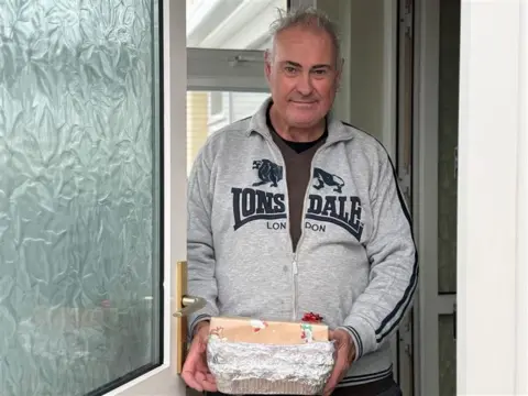 A man in a grey tracksuit top stands at his UPVC double-glazed front door holding a package.