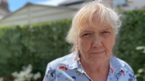 Sue Robertson, a woman with grey-white hair wearing a blue short-sleeved shirt with a large floral pattern